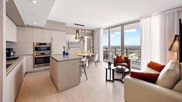 A modern apartment with a kitchen, dining area, and living room. It features a large window with a city view, a sofa, and a coffee table.