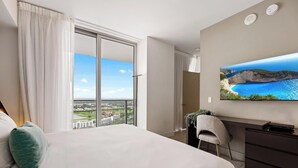 A hotel room with a large bed, a desk, a chair, a TV, and a view of a coastal landscape.