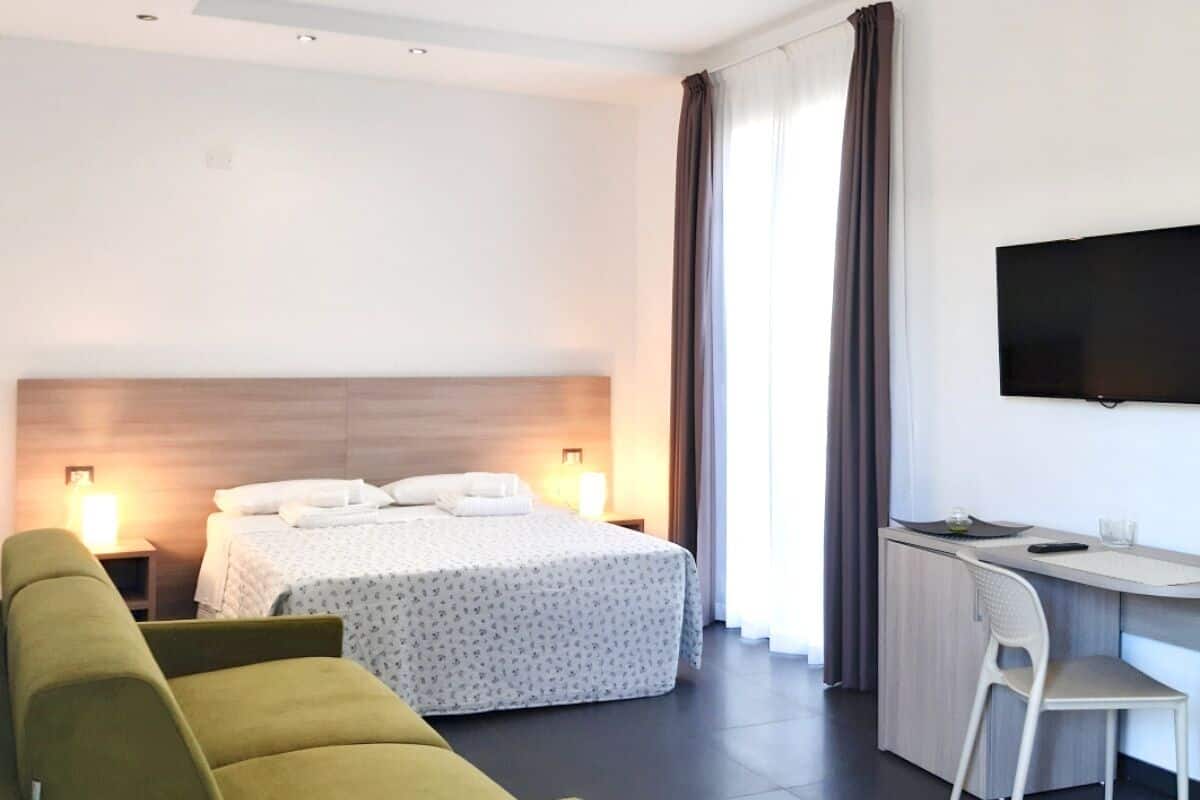 A modern hotel room with a bed, a green sofa, a desk with a chair, and a flat-screen TV.