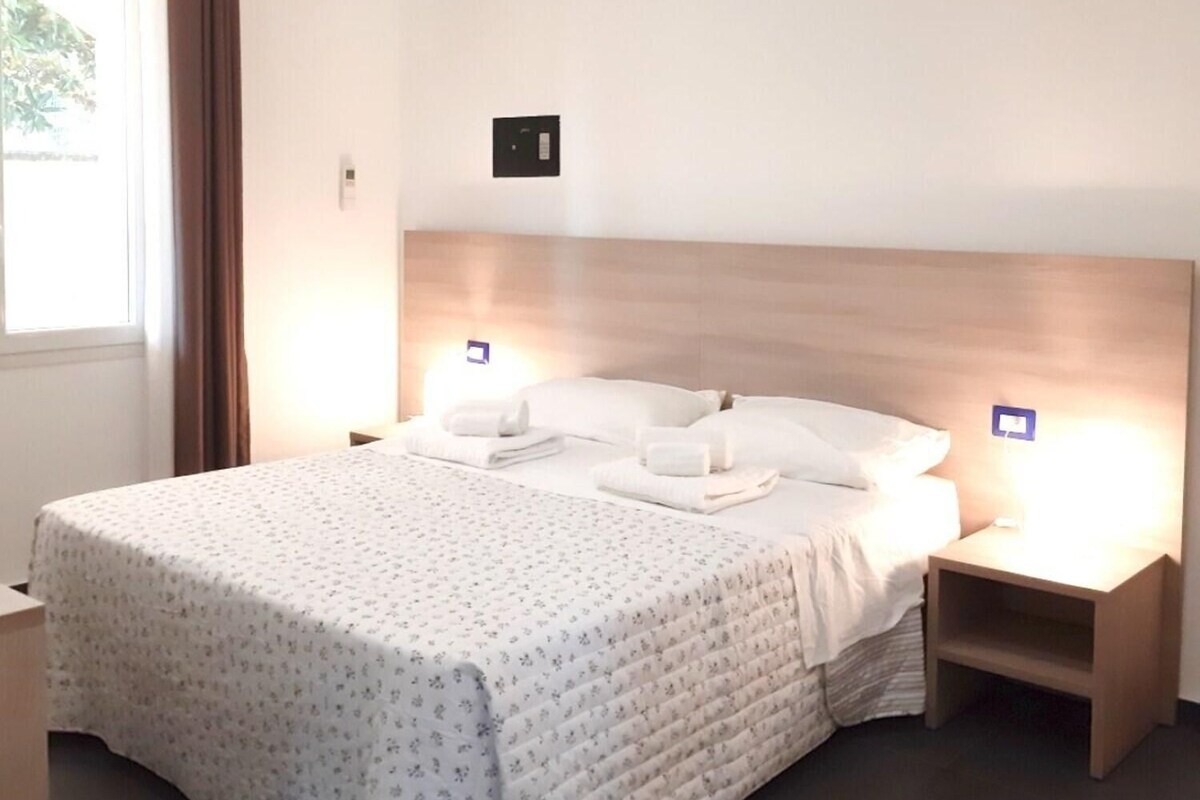 A neatly made double bed with a headboard, two bedside tables with lamps, and a window with curtains.