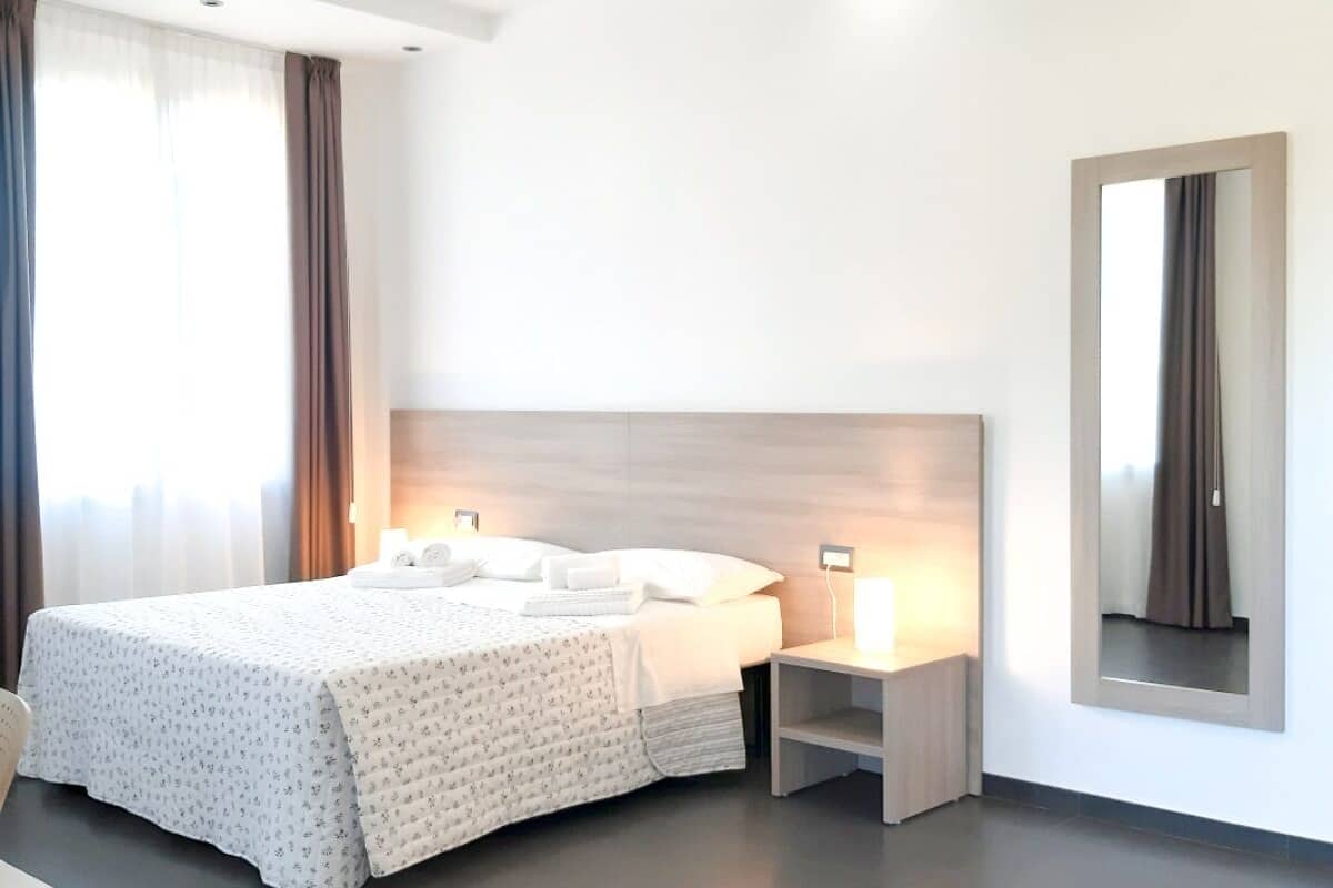 A modern hotel room with a large bed, bedside tables, a mirror, and a view of the outdoors.