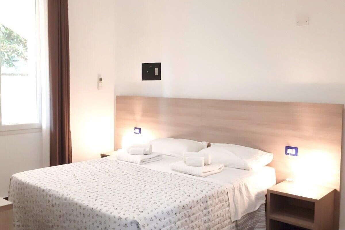 A hotel room with a large bed, two bedside tables, a window with curtains, and a wall-mounted light switch.