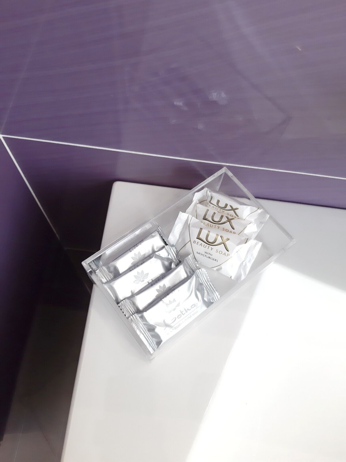 Ein Badezimmer mit einer violetten Wand und einer weißen Ablage. Darin ein durchsichtiger Kunststoffbehälter mit einer Lux Beauty Seife mit Feuchtigkeitszufuhr.