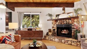 A cozy living room with a stone fireplace, a wooden ceiling, and a large window with curtains.