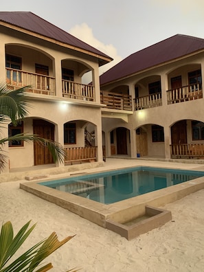 A two-story building with a swimming pool, multiple balconies, and a sandy ground.