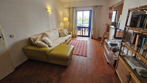A living room with a sofa, a small table, a bookshelf, and a view through a glass door to an outdoor area. - Recreational Vehicle (Vilamoura)