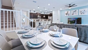 A modern dining area with a white table set for six, a kitchen in the background, and a staircase leading to an upper level.