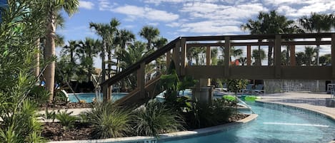 A swimming pool with a wooden bridge, surrounded by palm trees and a clear blue sky.