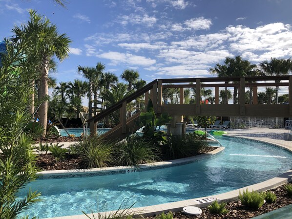 A swimming pool with a wooden bridge, surrounded by palm trees and a clear blue sky.