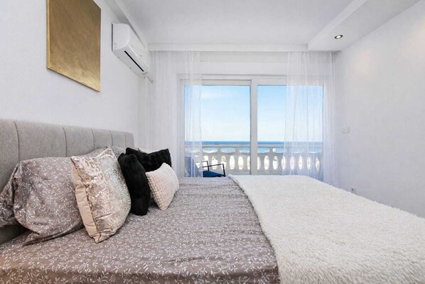 A bedroom with a bed, a sofa, a balcony with a view, and an air conditioning unit.