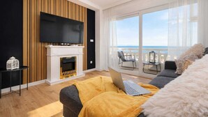 Una sala de estar moderna con chimenea, un televisor de pantalla plana, un balcón con vistas al mar y una computadora portátil sobre un sofá.