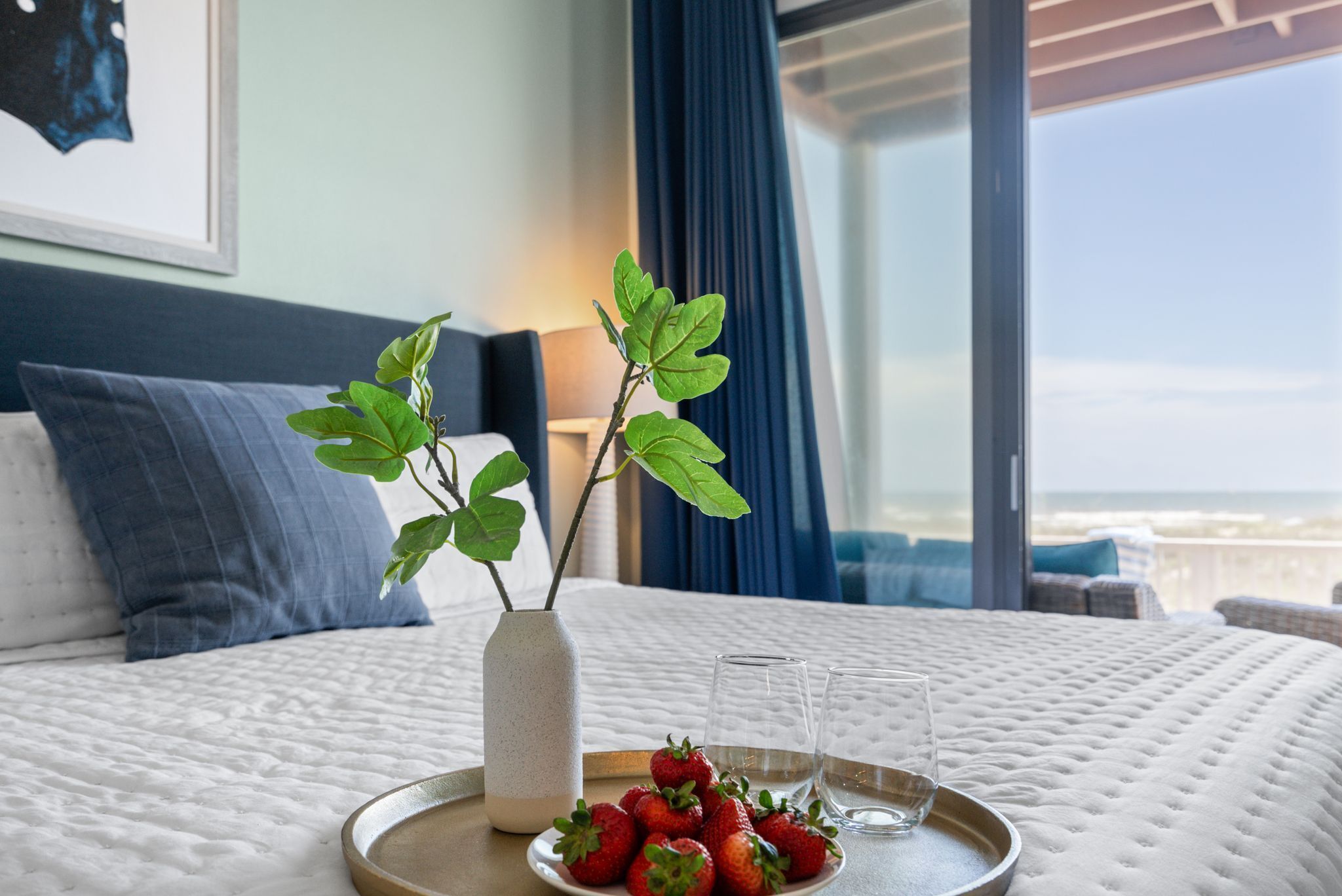 Een bed met een wit geborduurde spreideken, een dienblad met aardbeien en glazen, een vaas met een plant en uitzicht op de oceaan door een raam.