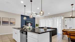 A modern kitchen with a central island, white cabinetry, and a dishwasher. There are pendant lights above the island and a dining area with a table and chairs. - Townhome with Star Wars Game Room, Private Pool, PS5 & Themed Bedrooms (Kissimmee)