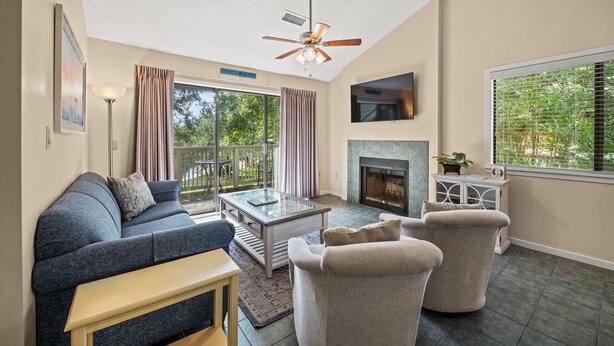 A spacious living room with a ceiling fan, a fireplace, a TV mounted on the wall, and a large window with blinds.