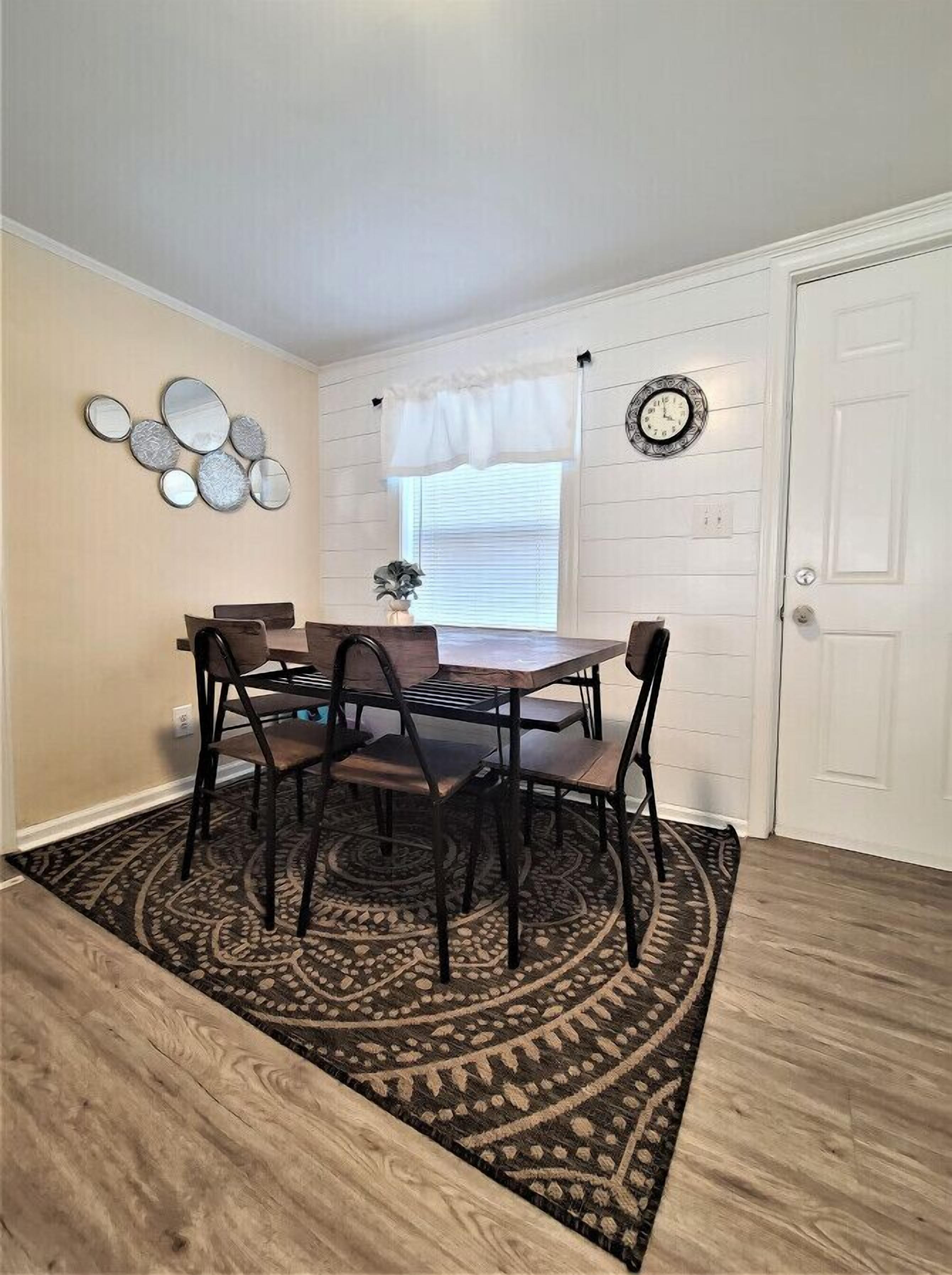 A dining area with a round table, four chairs, a patterned rug, a wall clock, and a window with blinds.