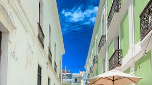 Un callejón empedrado con edificios a ambos lados, mesas al aire libre con sombrillas y un cielo azul despejado.