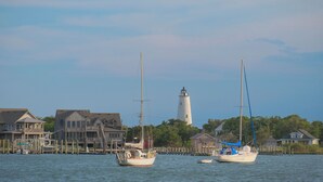 A waterfront scene with two sailboats docked near houses and a lighthouse in the background. - Bluff Shoal Motel (Ocracoke)