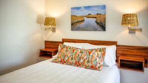 A neatly made bed with a white comforter and a colorful geometric pillow, flanked by two nightstands with woven lamps. A painting of a river landscape hangs above the bed. - Bluff Shoal Motel (Ocracoke)