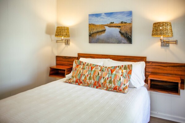 A neatly made bed with a white comforter and a colorful geometric pillow, flanked by two nightstands with woven lamps. A painting of a river landscape hangs above the bed. - Bluff Shoal Motel (Ocracoke)