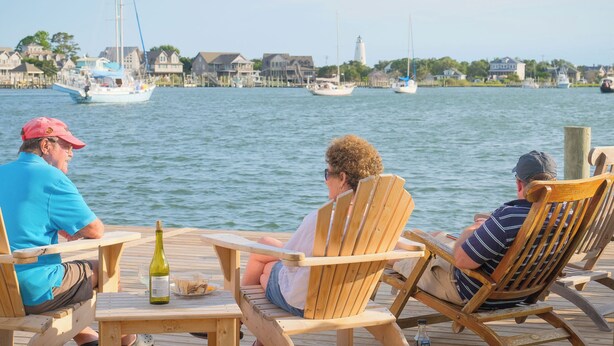 Three people sitting on wooden chairs on a dock, overlooking a marina with boats and houses in the background. - Bluff Shoal Motel (Ocracoke)