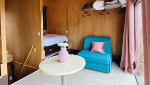 Una habitación compacta con una cama, un sofá azul, una mesita con un florero rosa y un ventilador de techo.