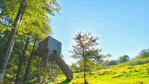 A rustic wooden structure with a sloped roof, elevated on stilts, surrounded by lush greenery and trees.