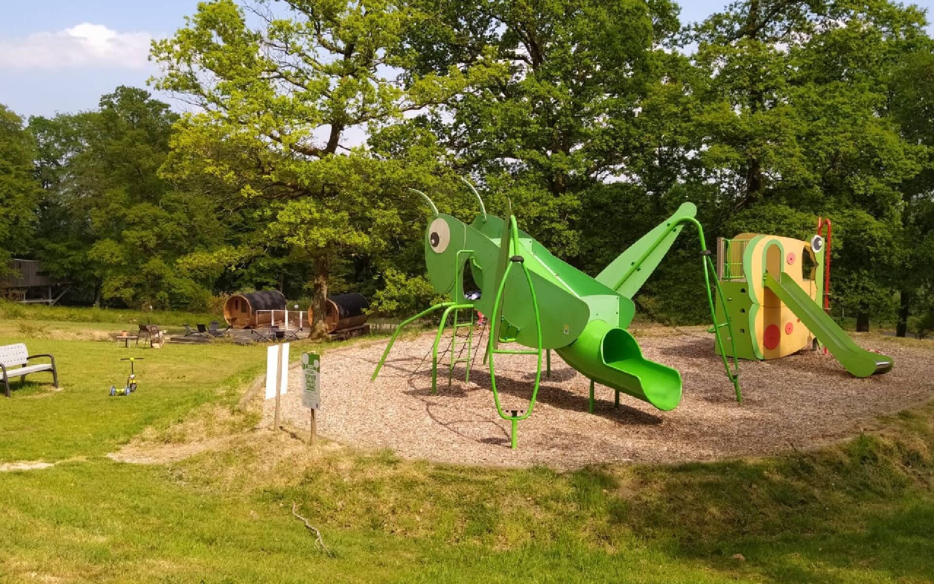 Un parque infantil con un tobogán verde y estructuras para escalar, rodeado de árboles y una zona de picnic.