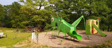 Un parque infantil con un tobogán verde y estructuras para escalar, rodeado de árboles y una zona de picnic.