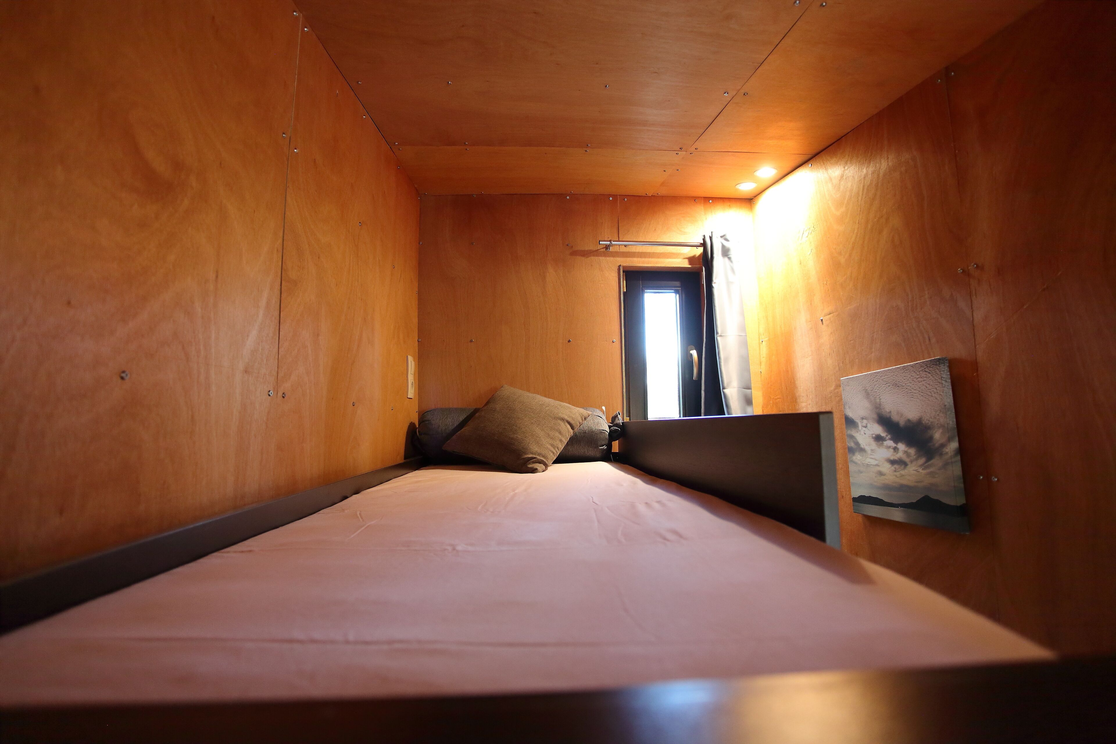 Ein Schlafzimmer mit Holzboden, einem Bett mit Matratze und Kissen, einem Nachttisch, einem Bild an der Wand und einem Fenster mit Vorhängen.