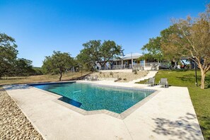 A rectangular swimming pool with clear blue water, surrounded by a concrete deck. There are lounge chairs and a small patio area with a stone wall.