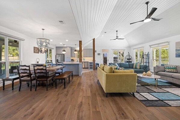 A spacious living area with a wooden floor, a yellow sofa, a dining table with chairs, and a kitchen in the background.