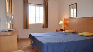 A hotel room with two beds, a wooden dresser, a desk with a lamp, a window with curtains, and a mirror on the wall. - Altura T1 Algarve Sun + Beach (Altura)