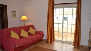 A red sofa with yellow cushions, a lamp, and a view through a glass door to an outdoor area. - Altura T1 Algarve Sun + Beach (Altura)