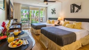 Une chambre d’hôtel avec deux lits, un ventilateur de plafond, une table à manger avec des fruits et des boissons, et un balcon offrant une vue sur la verdure.