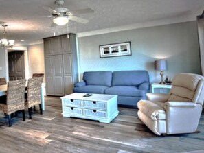 A living room with a ceiling fan, a blue sofa, a beige armchair, a white coffee table, and a dining area with chairs.