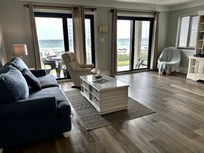 A living room with a sofa, armchair, coffee table, and a view of the beach through large sliding doors.