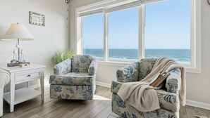 Une pièce dotée d’une grande fenêtre donnant sur l’océan, de deux fauteuils à motifs, d’un bureau blanc et d’une lampe.