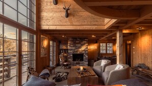 A cozy cabin interior with a stone fireplace, wooden beams, and a large window overlooking a deck.
