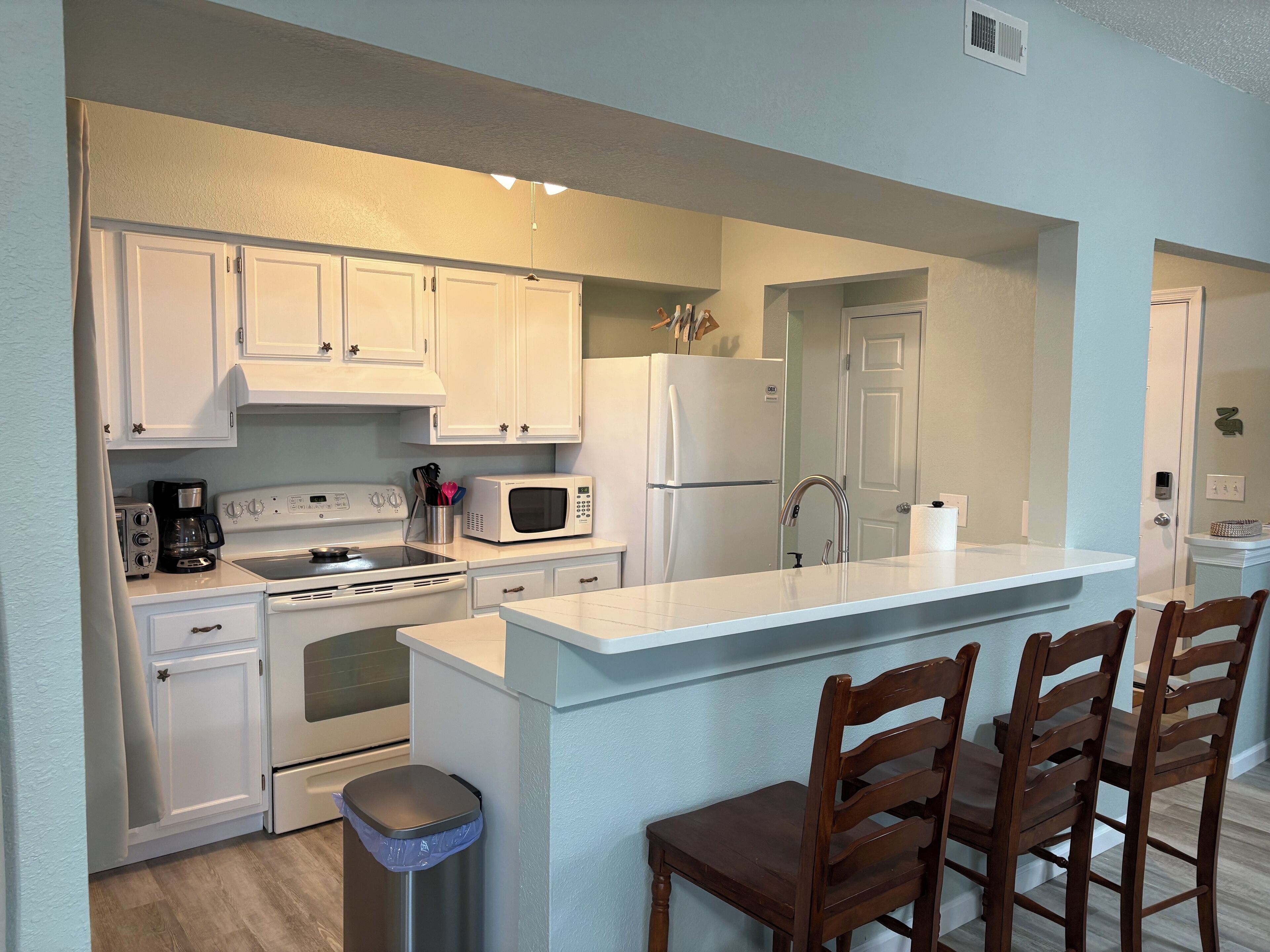 A modern kitchen with white cabinets, a stainless steel sink, a built-in microwave, and a refrigerator. There are wooden chairs at the island and a trash can.