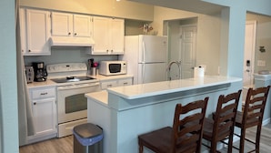 A modern kitchen with white cabinets, a stainless steel sink, a built-in microwave, and a refrigerator. There are wooden chairs at the island and a trash can. - Newly remodeled. Steps away from the ocean! Condo in Kill Devil Hills. (Kill Devil Hills)