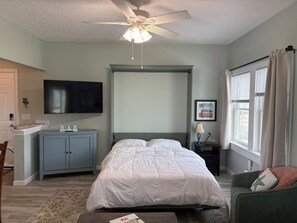Quarto com cama, escrivaninha, televisão, ventoinha no teto e janela com cortinas.