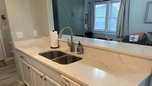 A kitchen with a double sink, white cabinets, and a dishwasher. - Newly remodeled. Steps away from the ocean! Condo in Kill Devil Hills. (Kill Devil Hills)