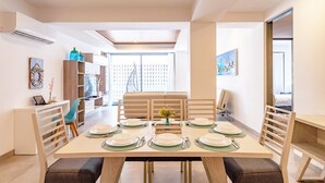 Ein moderner Essbereich mit einem großen Tisch, gedeckt für sechs Personen, umgeben von Holzstühlen. Es gibt eine Sitzecke mit einer Couch und einem Hängekorbsessel. Der Raum hat ein großes Fenster mit Blick auf einen Zäun mit Musterung.