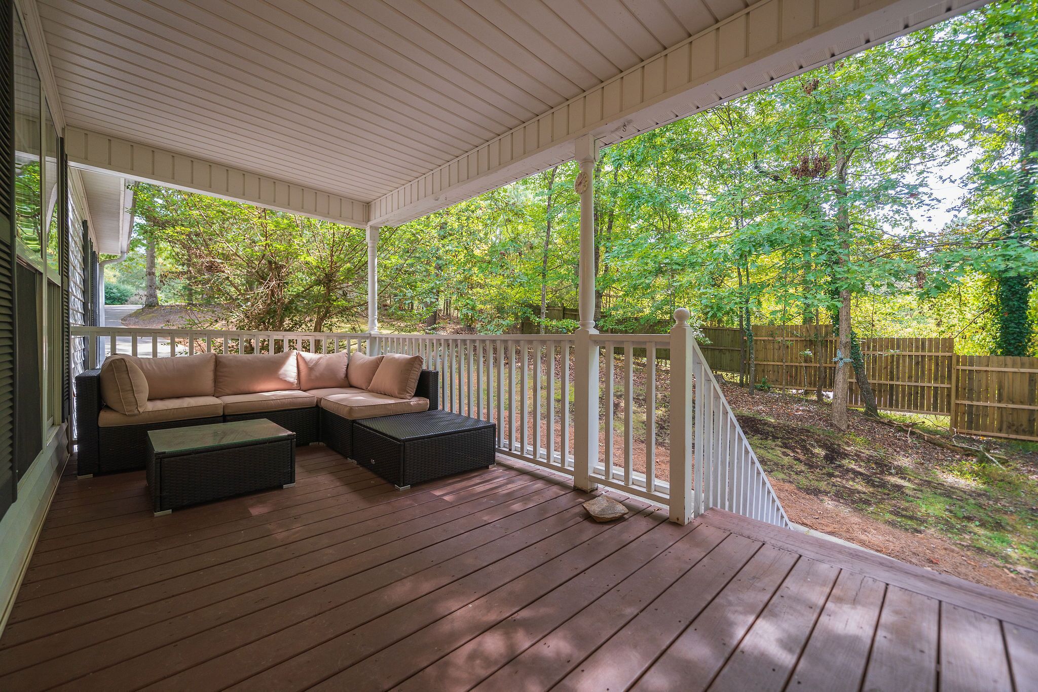 A covered porch with a beige sofa and two black ottomans, a wooden deck, and a view of a wooded area.