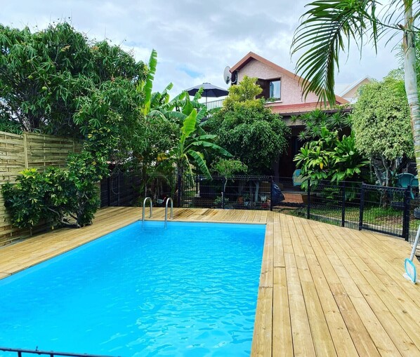 A rectangular swimming pool with a wooden deck surrounding it, set against a backdrop of lush greenery and a house. - Villa Sea View (Saint-Leu)