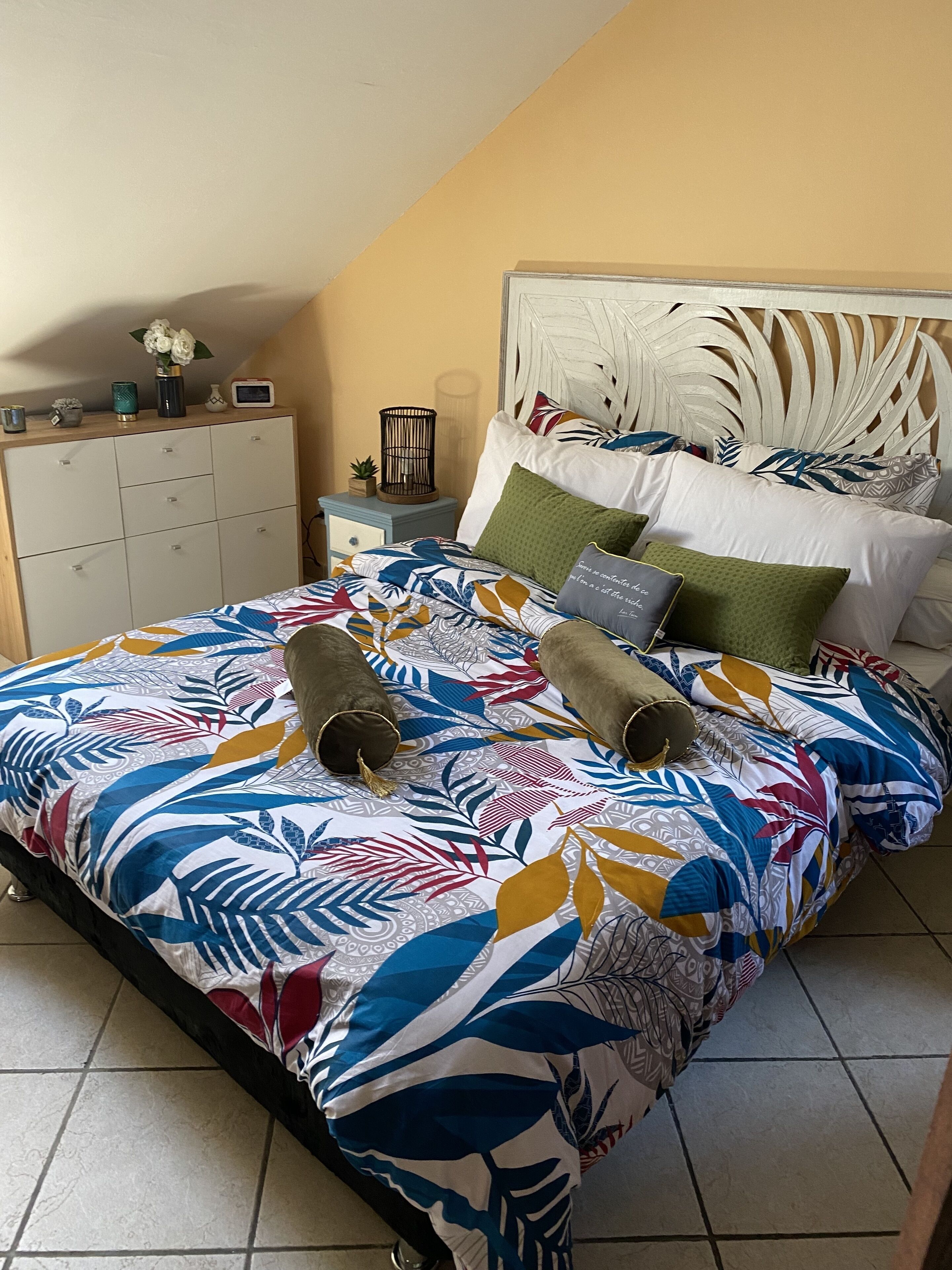 A bedroom with a bed featuring a colorful leaf-patterned bedspread, a wooden headboard, and multiple pillows. There is a nightstand with a lamp and a small plant.