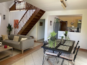 Ein Wohnzimmer mit grauem Sofa, einem Esstisch mit Stühlen, einer Treppe und einem Küchenbereich im Hintergrund.