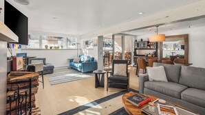 Amplia sala de estar con sofá, sillones y una mesa de centro. Se observa una chimenea, un comedor y una cocina al fondo.