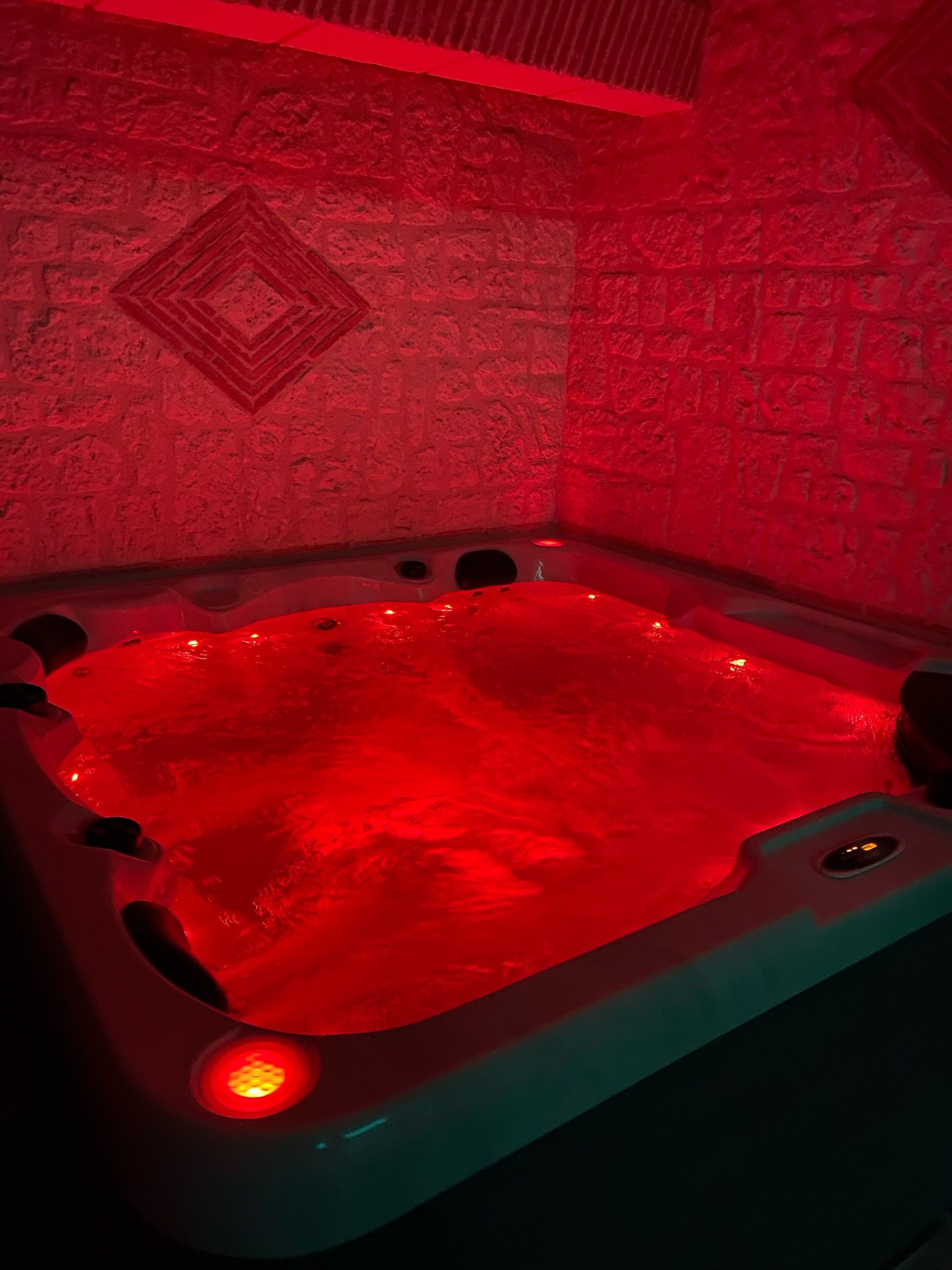 Un bain à remous éclairé d’une lumière rouge, placé devant un mur texturé orné d’un motif géométrique.