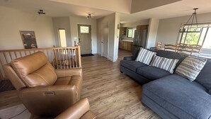 A spacious living room with a leather chair, a gray sofa with striped pillows, wooden flooring, and a view of the kitchen and dining area.